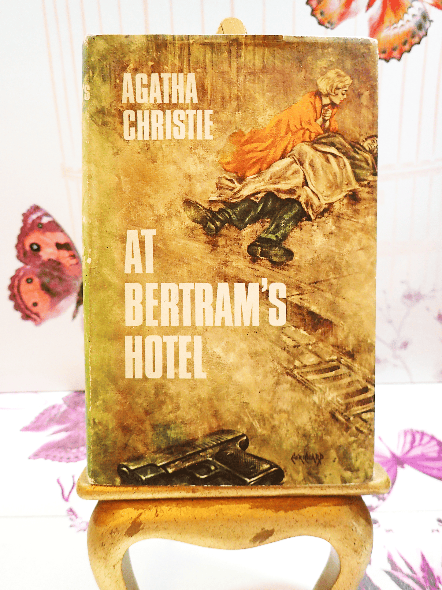Front cover of Agatha Christie's At Bertrams Hotel Vintage Book Classic Crime Fiction 1950's Hardback showing a man lying on the street with a woman attending to him and a gun at the forefront.
