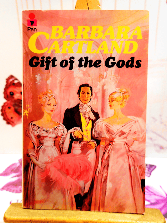Front cover of Gift of the Gods Barbara Cartland Pan Paperback First Edition showing two pretty women dressed in pink Victorian Dresses in front of handsome man.