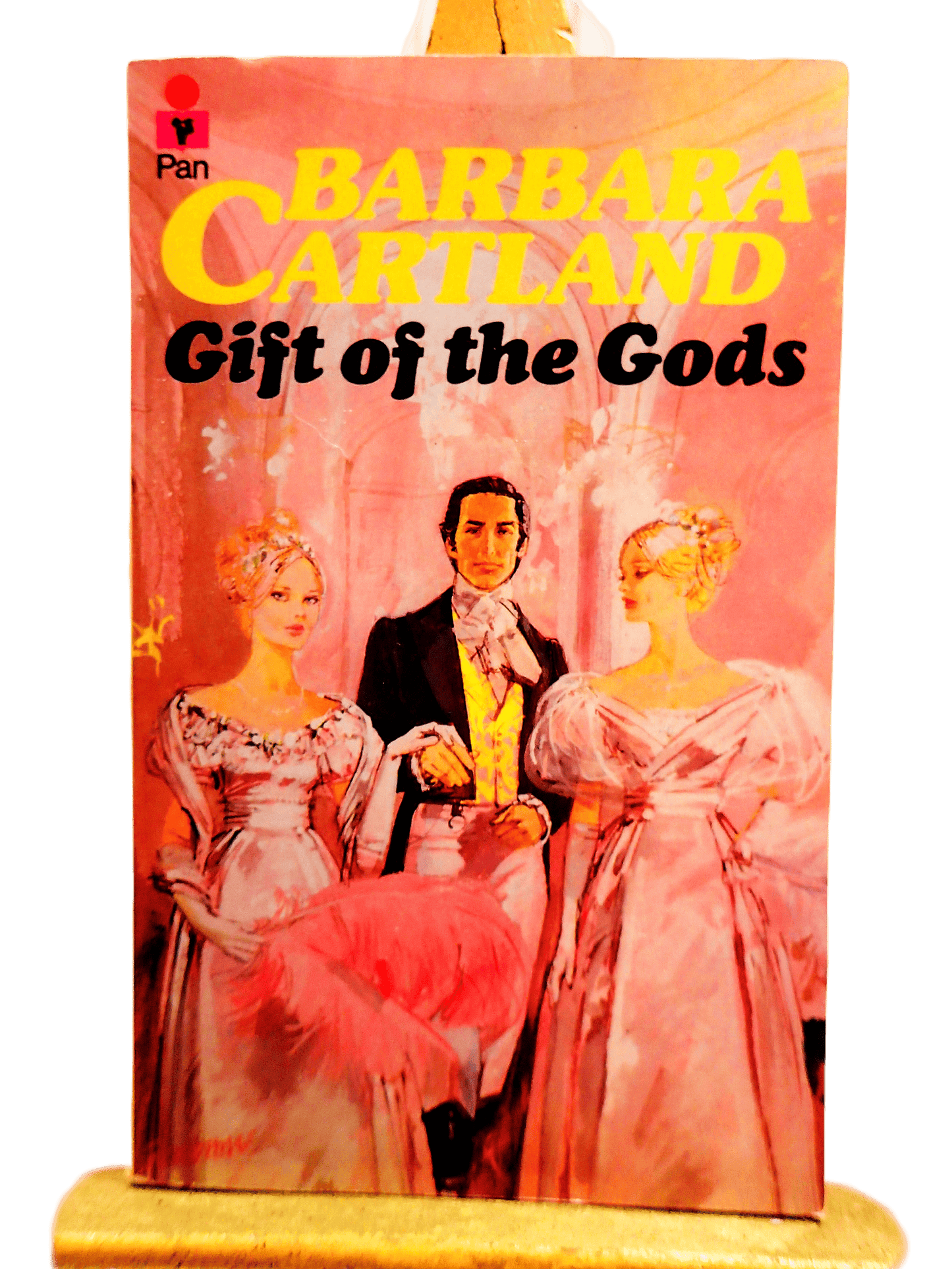 Front cover of Gift of the Gods Barbara Cartland Pan Paperback First Edition showing two pretty women dressed in pink Victorian Dresses in front of handsome man.