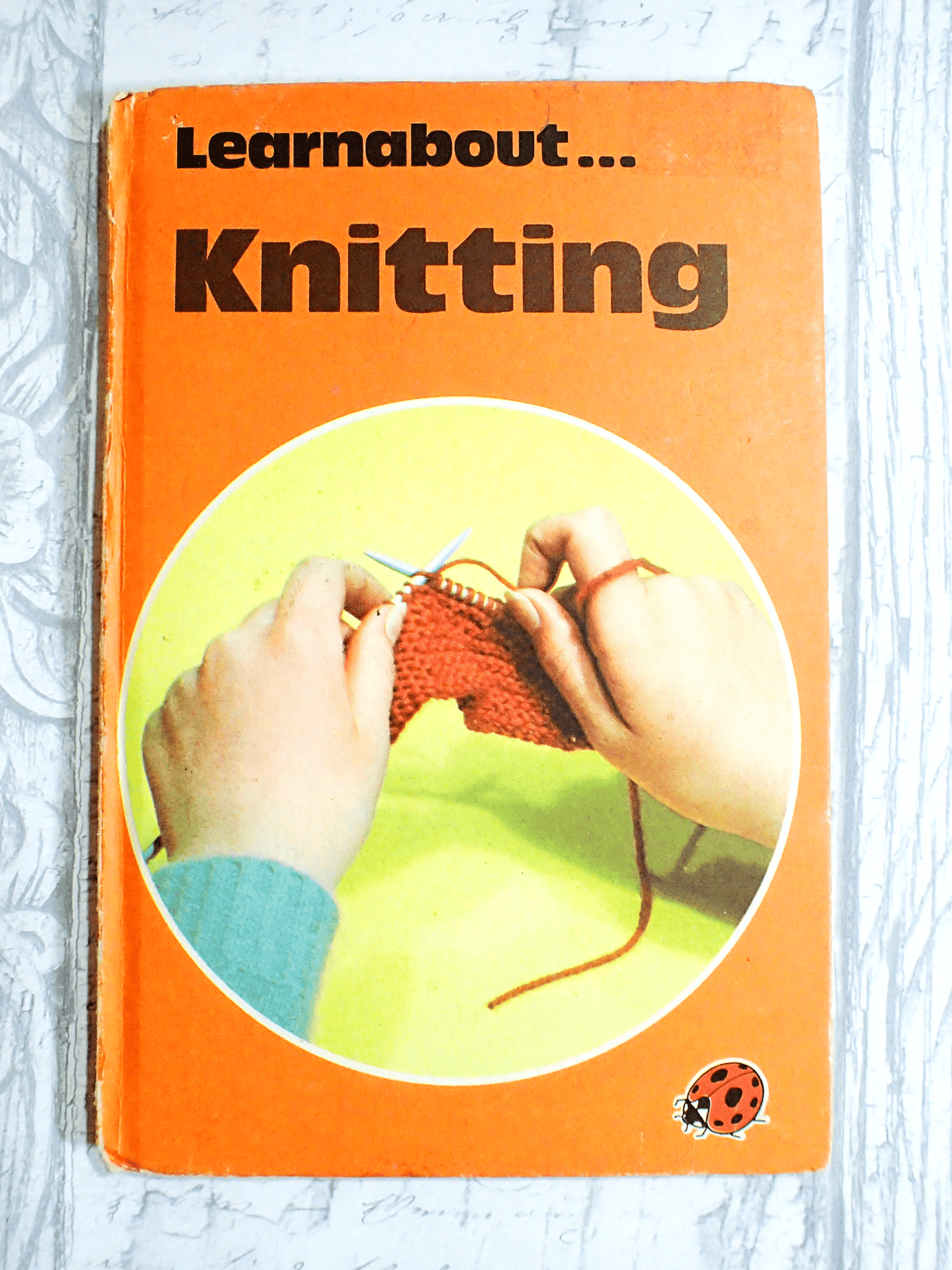 Learnabout knitting cover of vintage book by Ladybird with orange cover and hands knitting, against a light background.