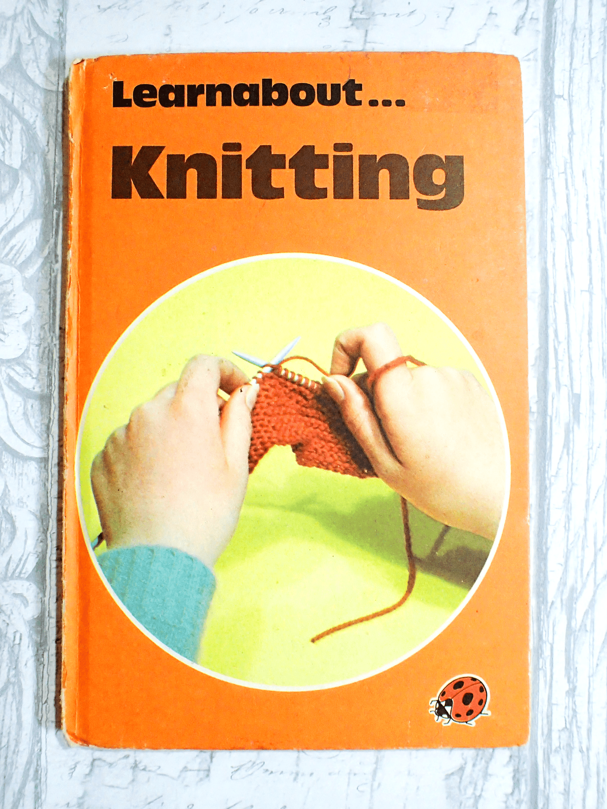 Learnabout knitting cover of vintage book by Ladybird with orange cover and hands knitting, against a light background.