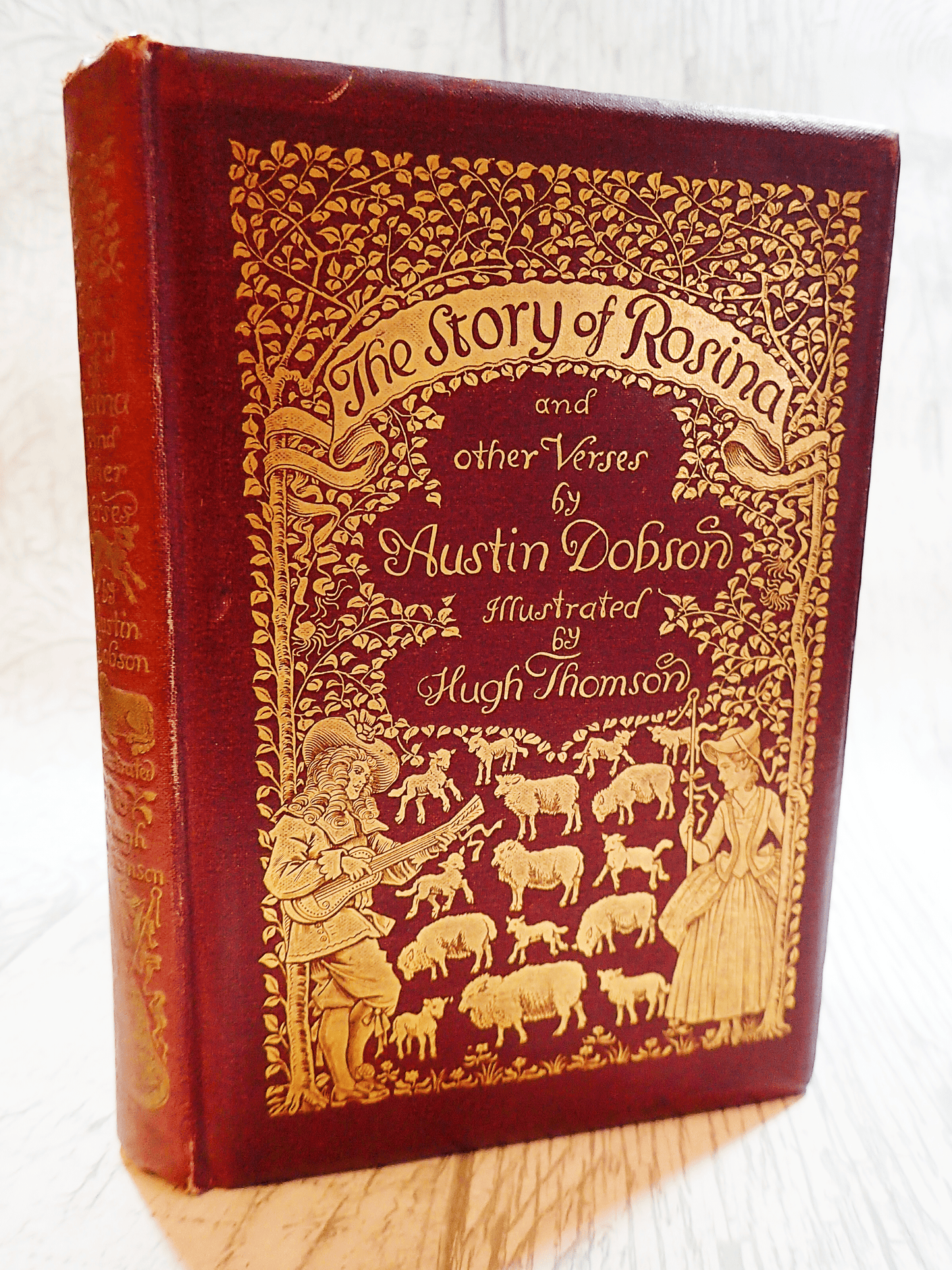 Ornate gilt embossed front cover of The Story of Rosina Austin Dobson illus Hugh Thompson First Edition Antique Book. Showing a french shepherdess with a lute player. Against a light background.
