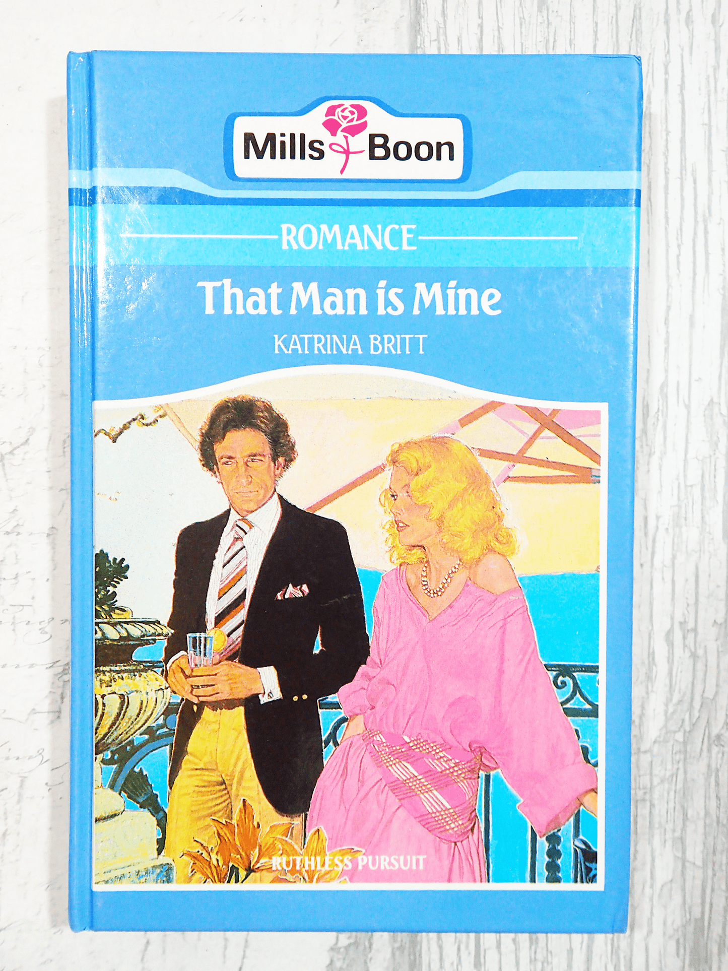 Front cover of Vintage Mills and Boon Romance That Man is Mine by Katrina Britt Hardback book 1980's showing A blonde woman and handsome man having a drink on a light background.