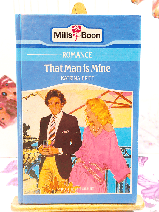 Front cover of Vintage Mills and Boon Romance That Man is Mine by Katrina Britt Hardback book 1980's showing A blonde woman and handsome man having a drink on a pastel blue ground. 
