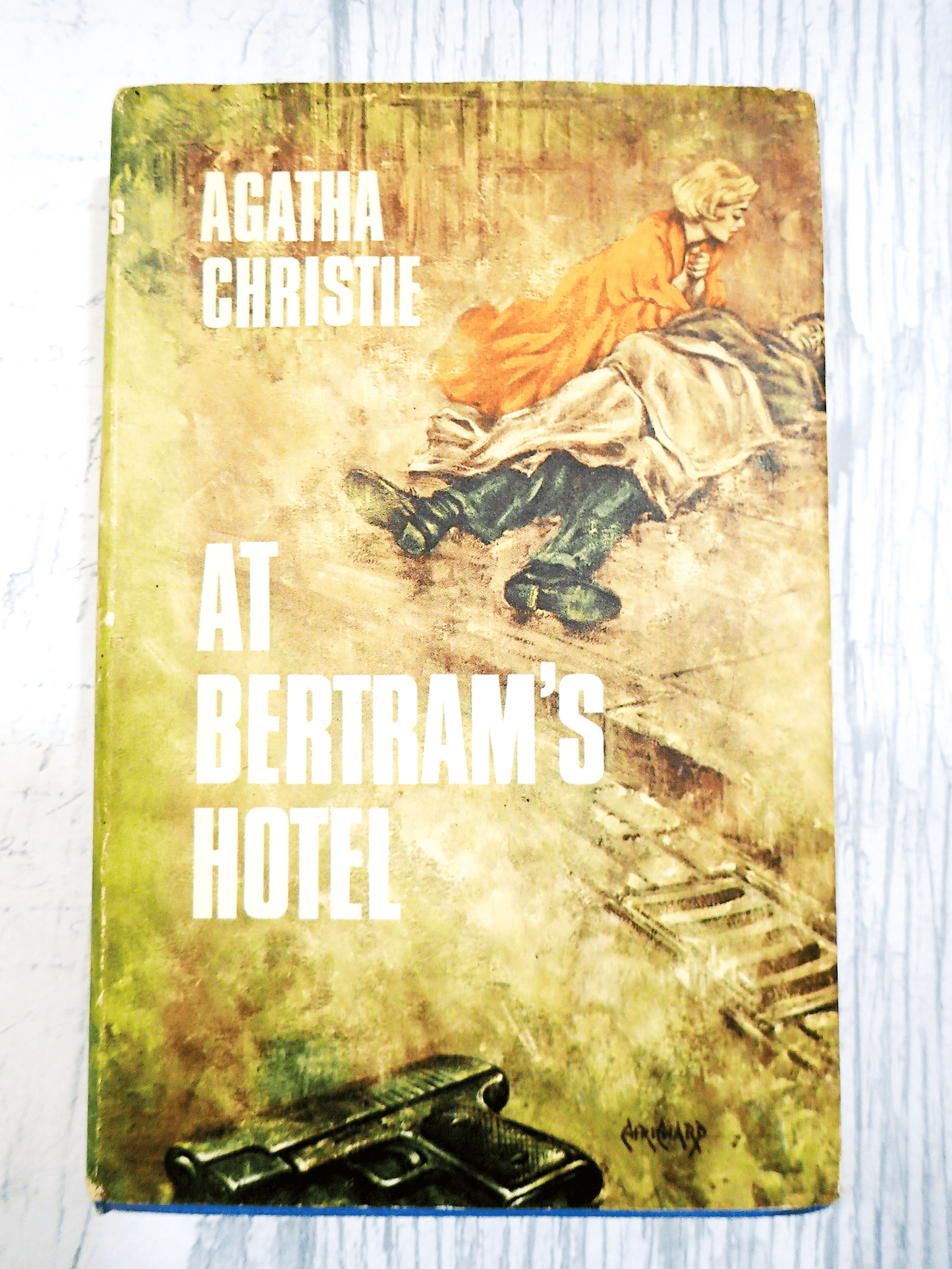 Front cover of Agatha Christie's At Bertrams Hotel Vintage Book Classic Crime Fiction 1950's Hardback showing a man lying on the street with a woman attending to him and a gun at the forefront against a light background. 