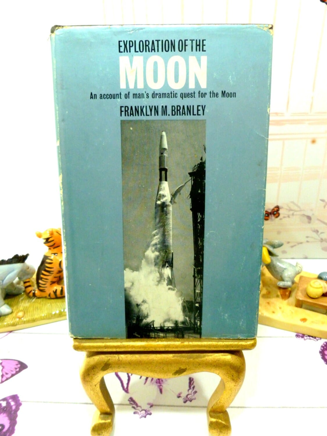Front cover of vintage book Exploration of the Moon showing a rocket launch against a metallic blue ground. 