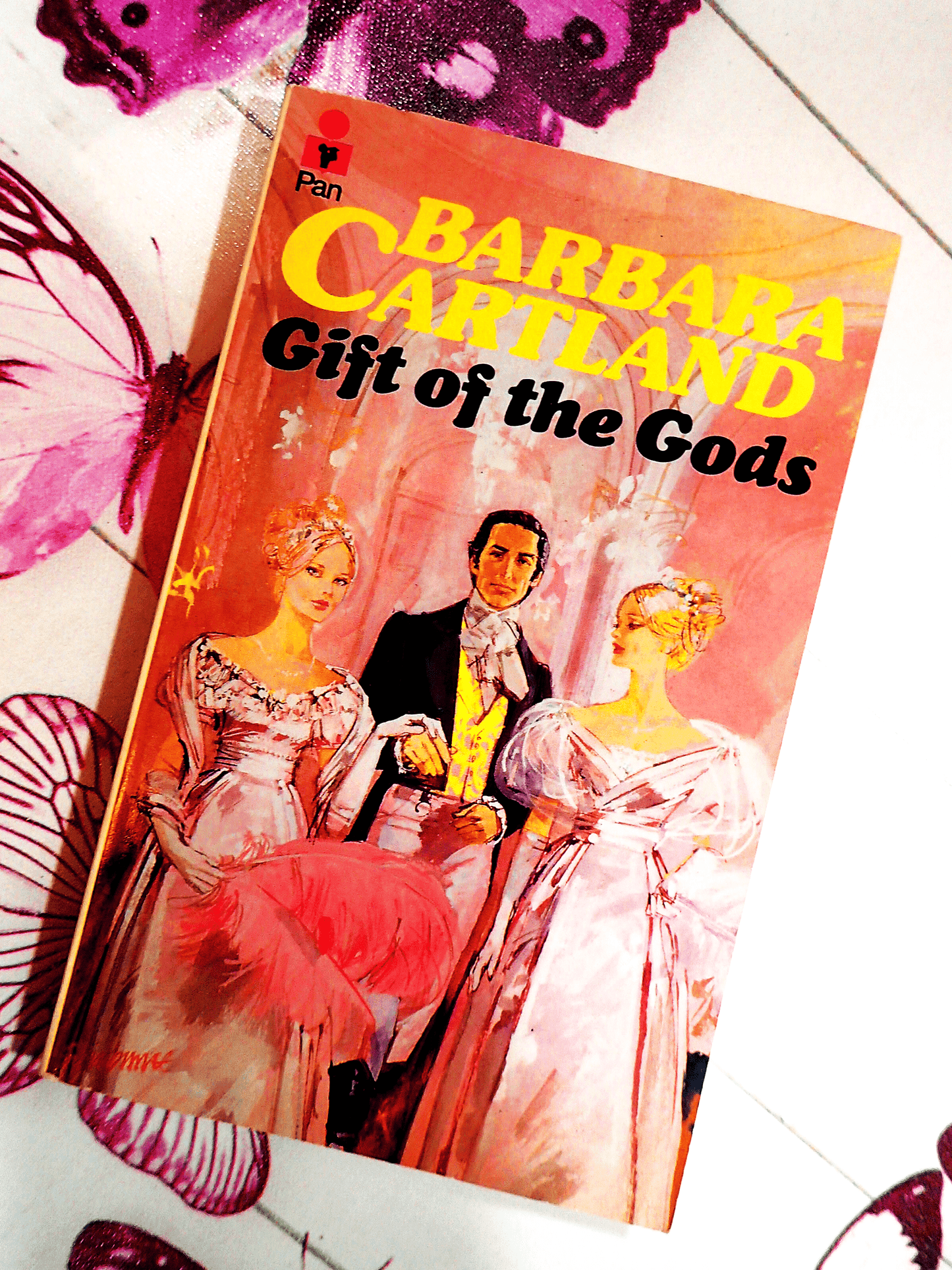 Front cover of Gift of the Gods Barbara Cartland Pan Paperback First Edition showing two pretty women dressed in pink Victorian Dresses in front of handsome man. 