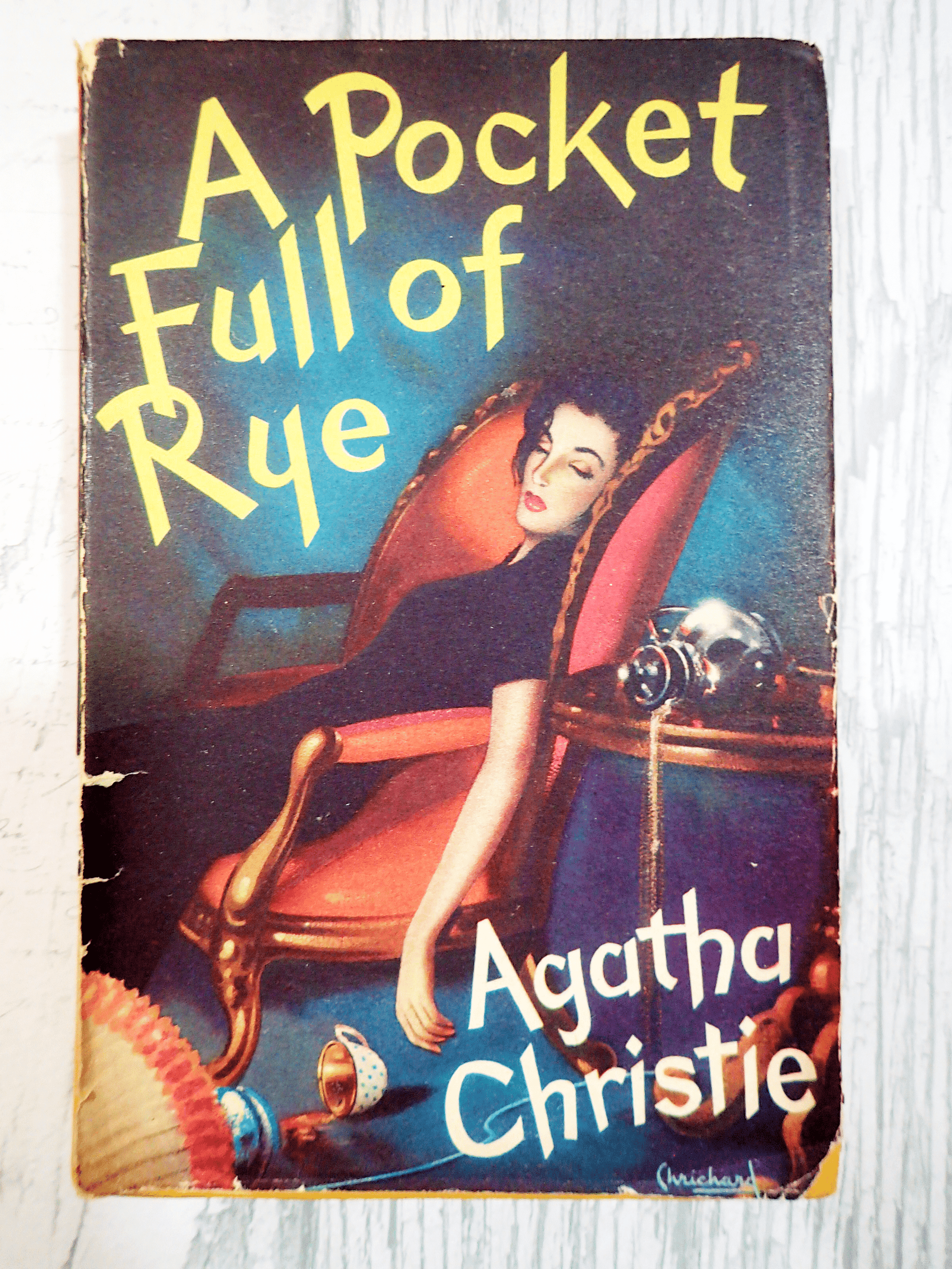 Front cover of A Pocketful of Rye Agatha Christie Vintage Thriller Book Club First Edition 1954 showing an attractive dark haired woman slumped in a chair with a coffee cup on the ground. Against a light background.