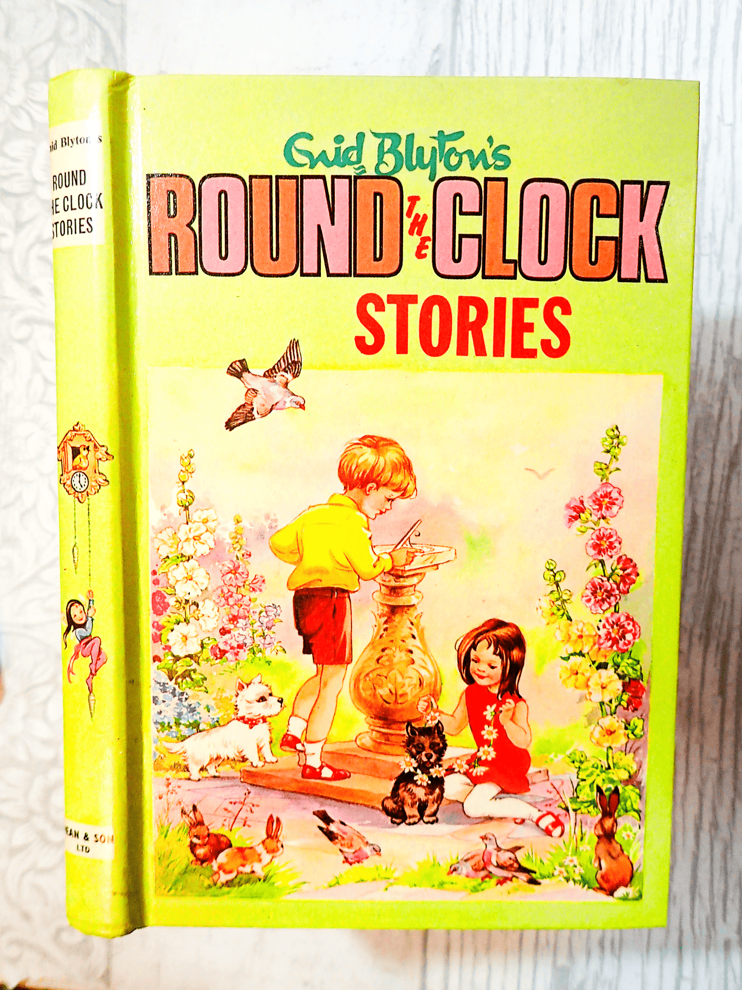 Front cover of Enid Blyton's Round the Clock Stories Vintage Children's Book 1970's Bedtime Fairytales showing children and dogs playing round a sundial against a light background 