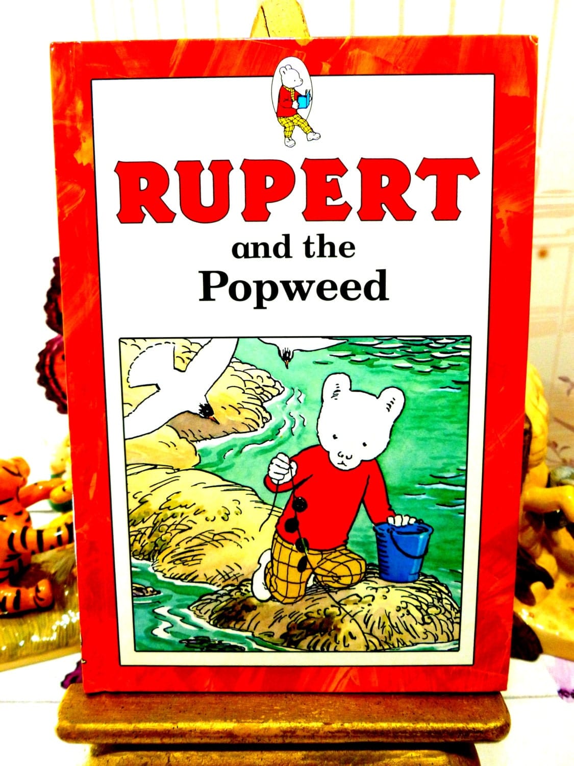 Front cover of Rupert and the Popweed Vintage Hardback Rupert Book 1st Ed 1991 showing Rupert and a Seagull against a red border. 